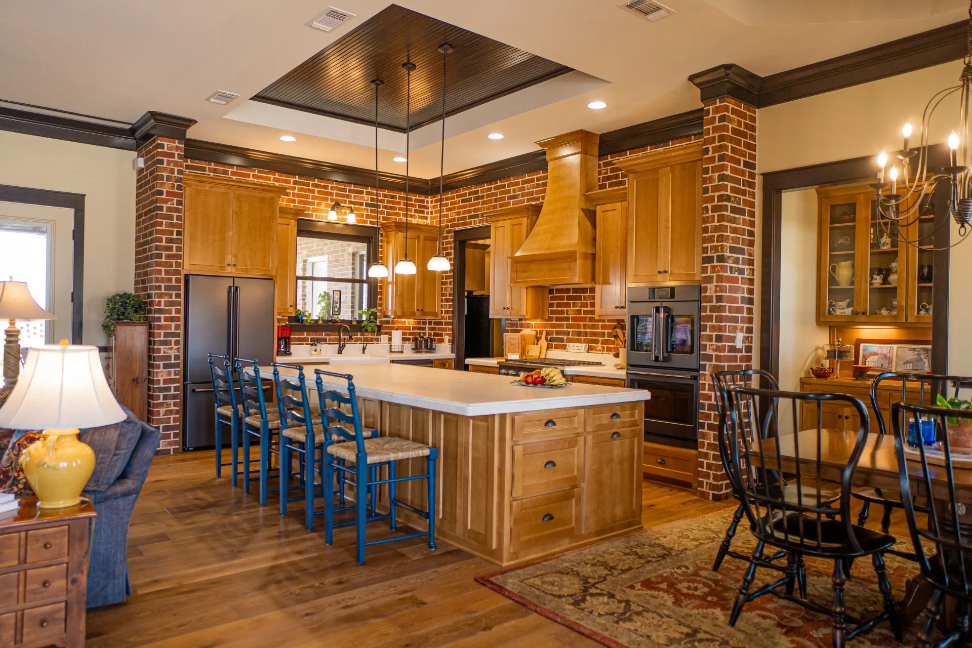 Open kitchen at the Wraparound Porch Home with a large wood island under a cream quartz top, honey-oak cabinetry and three pendant lights, with a brick dining alcove and an oriental rug beyond
