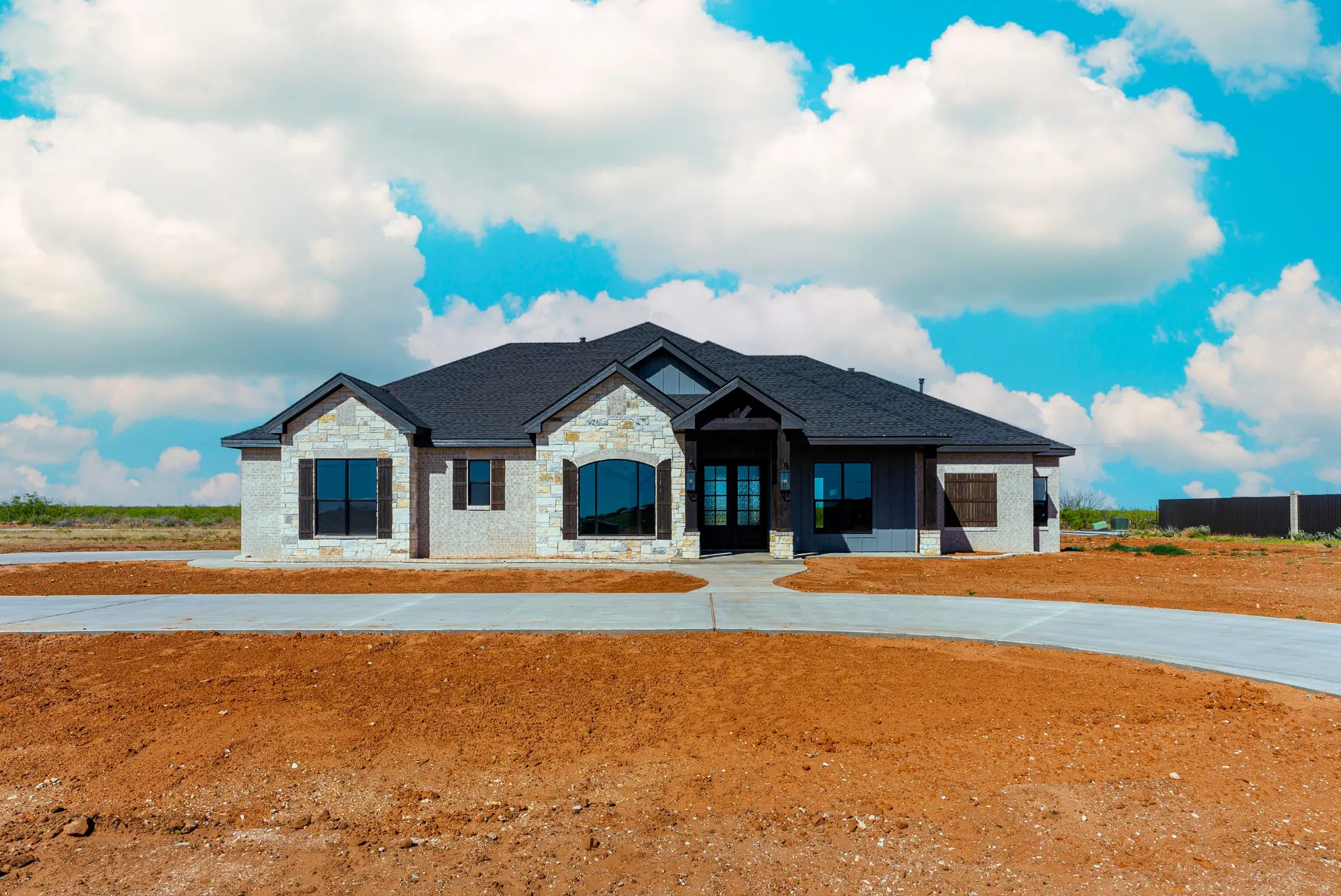 Front exterior of the Eliam St Home with stone and white stucco, a dark gabled roof, timber-framed entry porch and a curved concrete drive over fresh red dirt