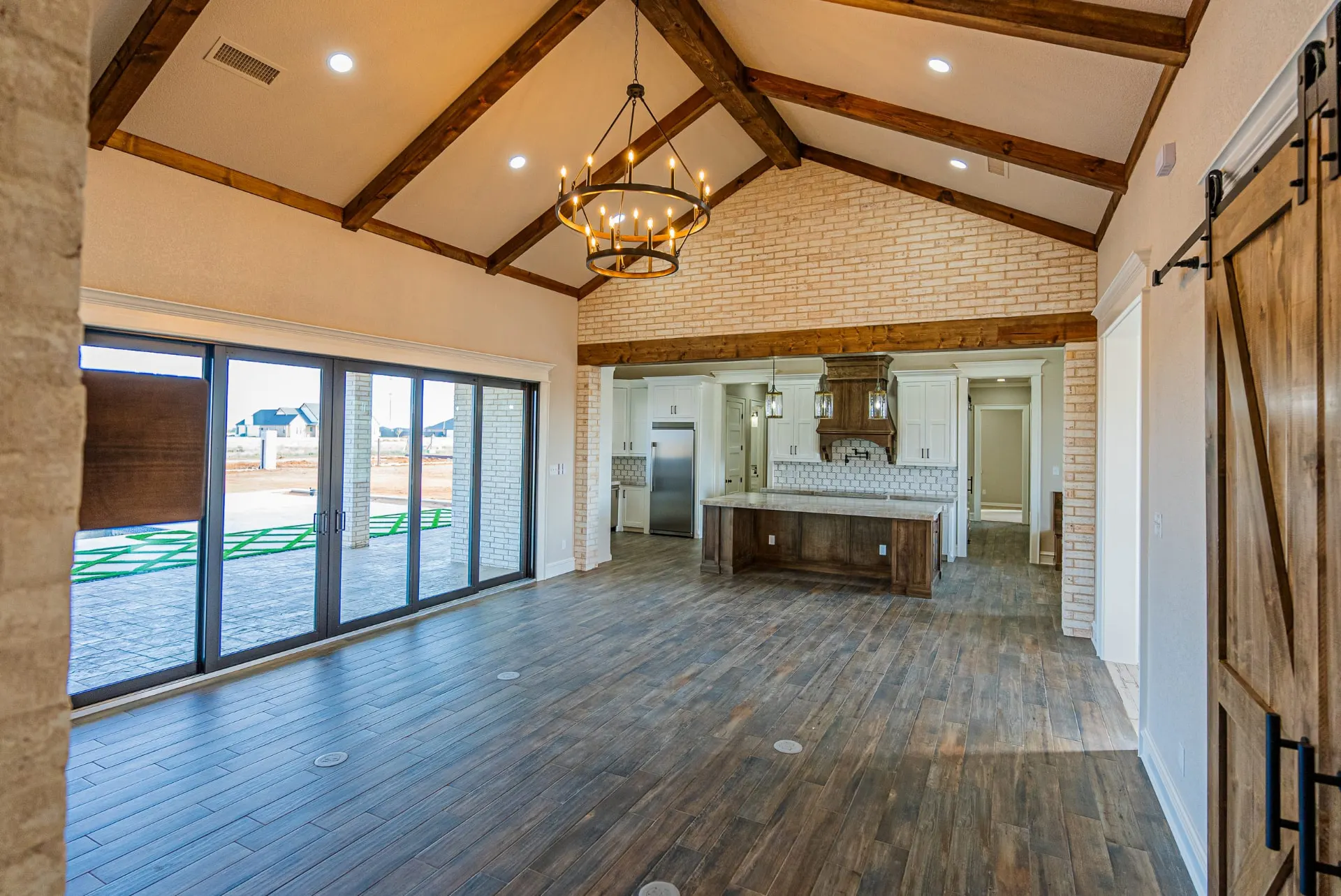 Vaulted great room at 1602 NW 12th St with exposed timber trusses, ring chandelier, whitewashed brick and a glass wall opening to the patio