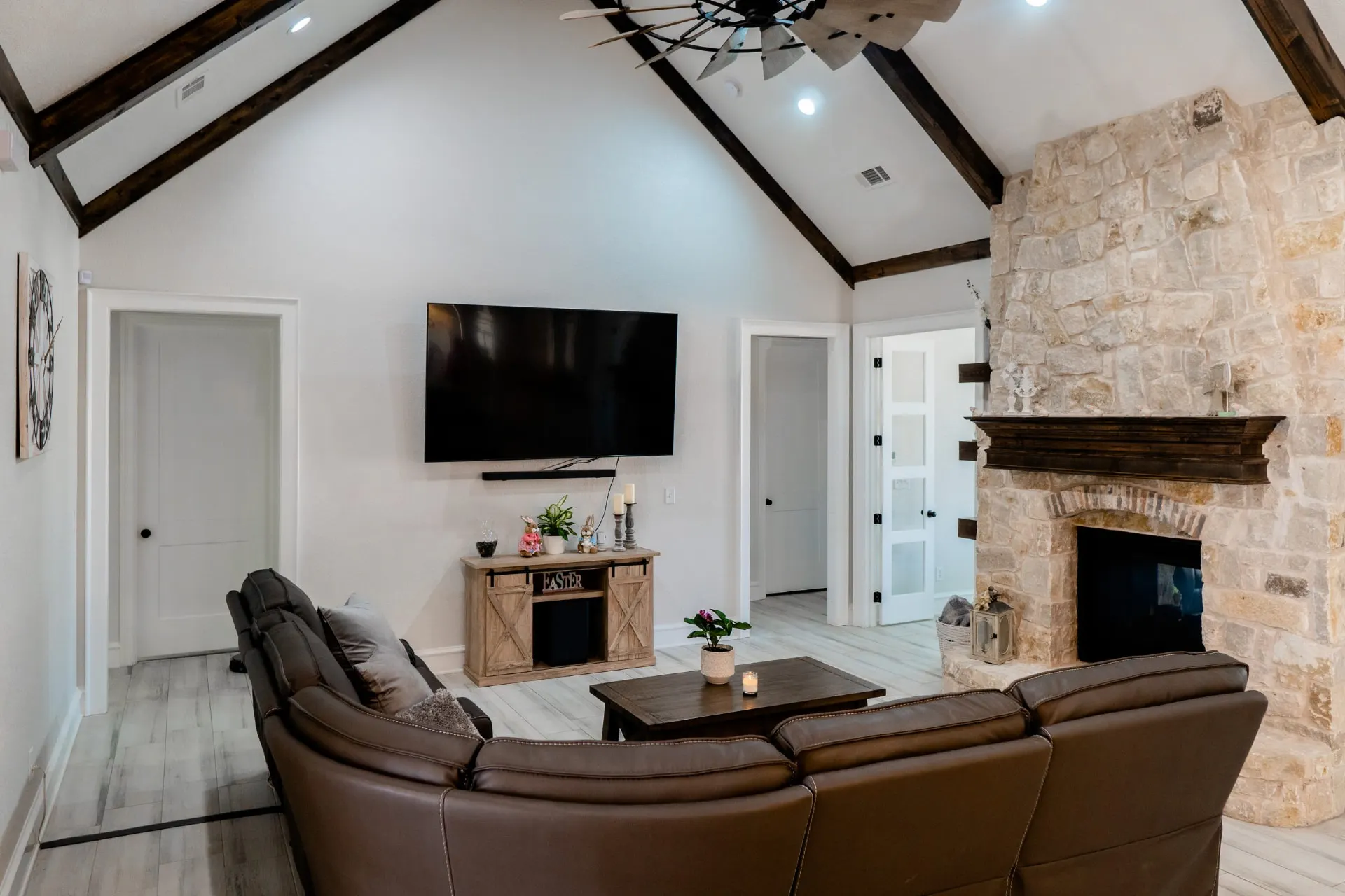 Great room at 1667 Eldenrod with vaulted dark-stained beams, a windmill-style fan, stone fireplace with mounted TV and leather seating