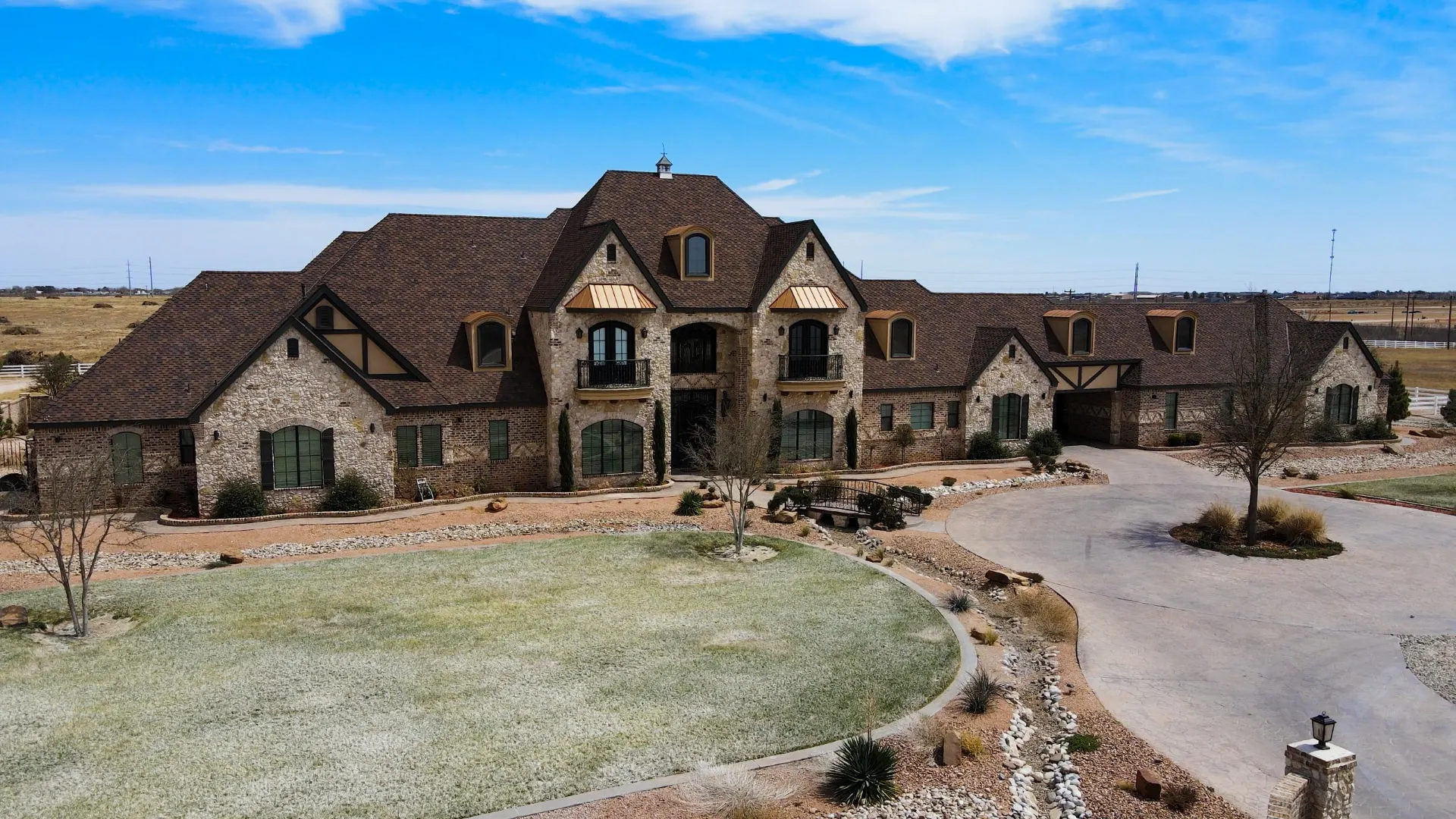 Aerial view of The Flores Estate — a stone French-country estate with steep multi-gabled roofs, eyebrow dormers, copper awnings, Juliet balconies and a covered breezeway, set on flat acreage outside Andrews, TX