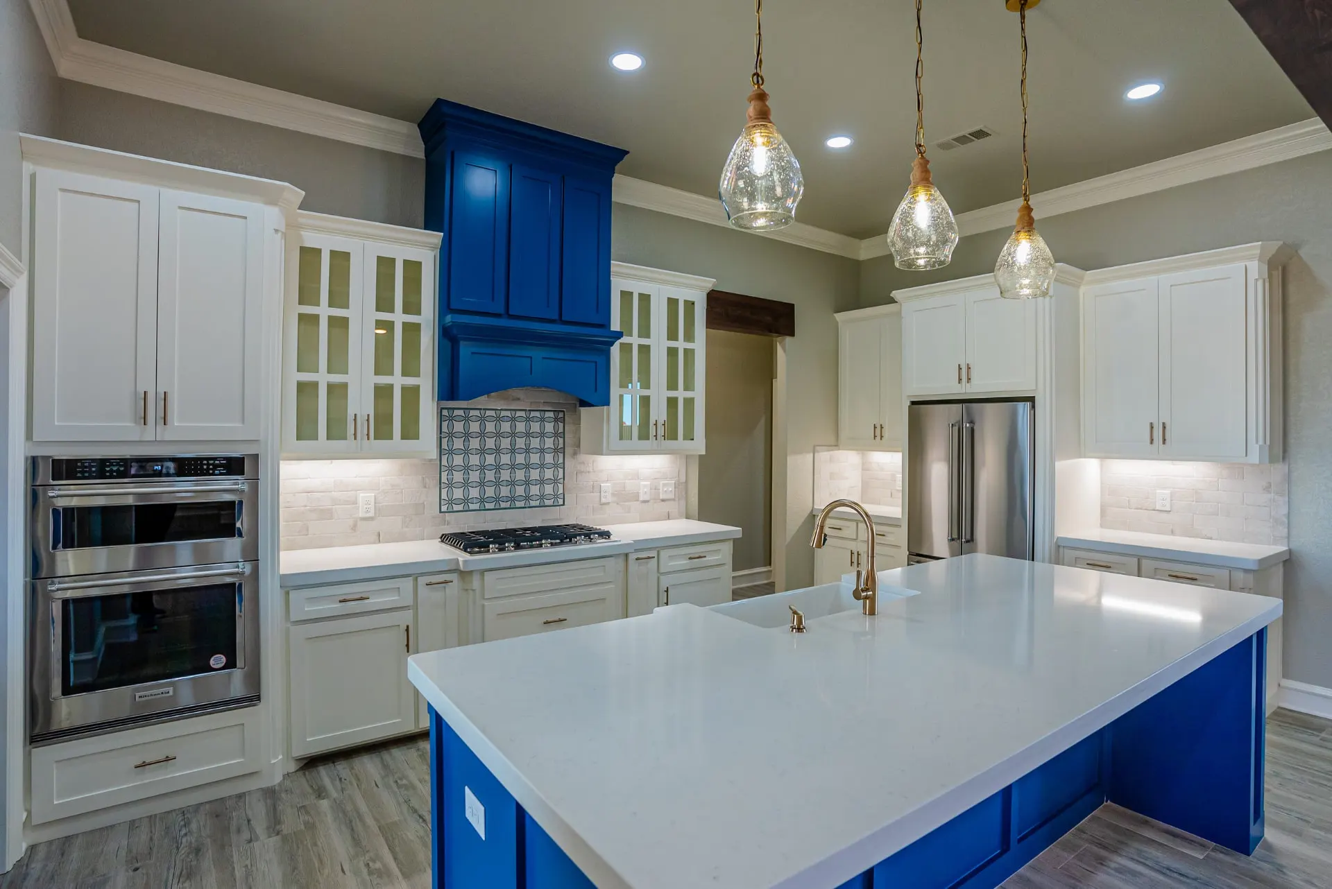 Eliam St Home kitchen viewed across the cobalt island toward the range hood, with double wall ovens and a stainless refrigerator beyond