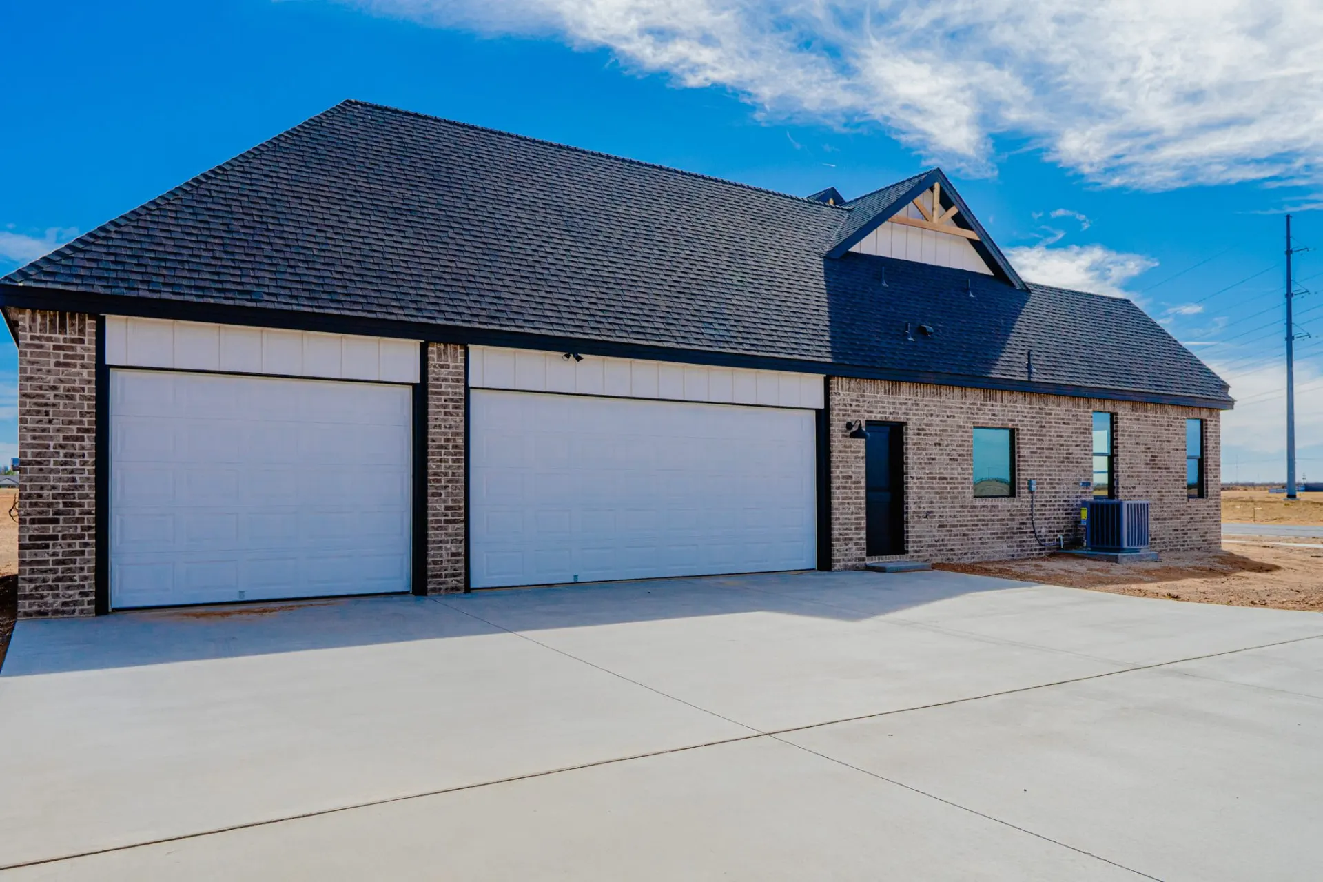 Exterior of the 12th St Home with grey brick, white board-and-batten over twin garage doors, a dark shingle roof and a timber-trimmed gable