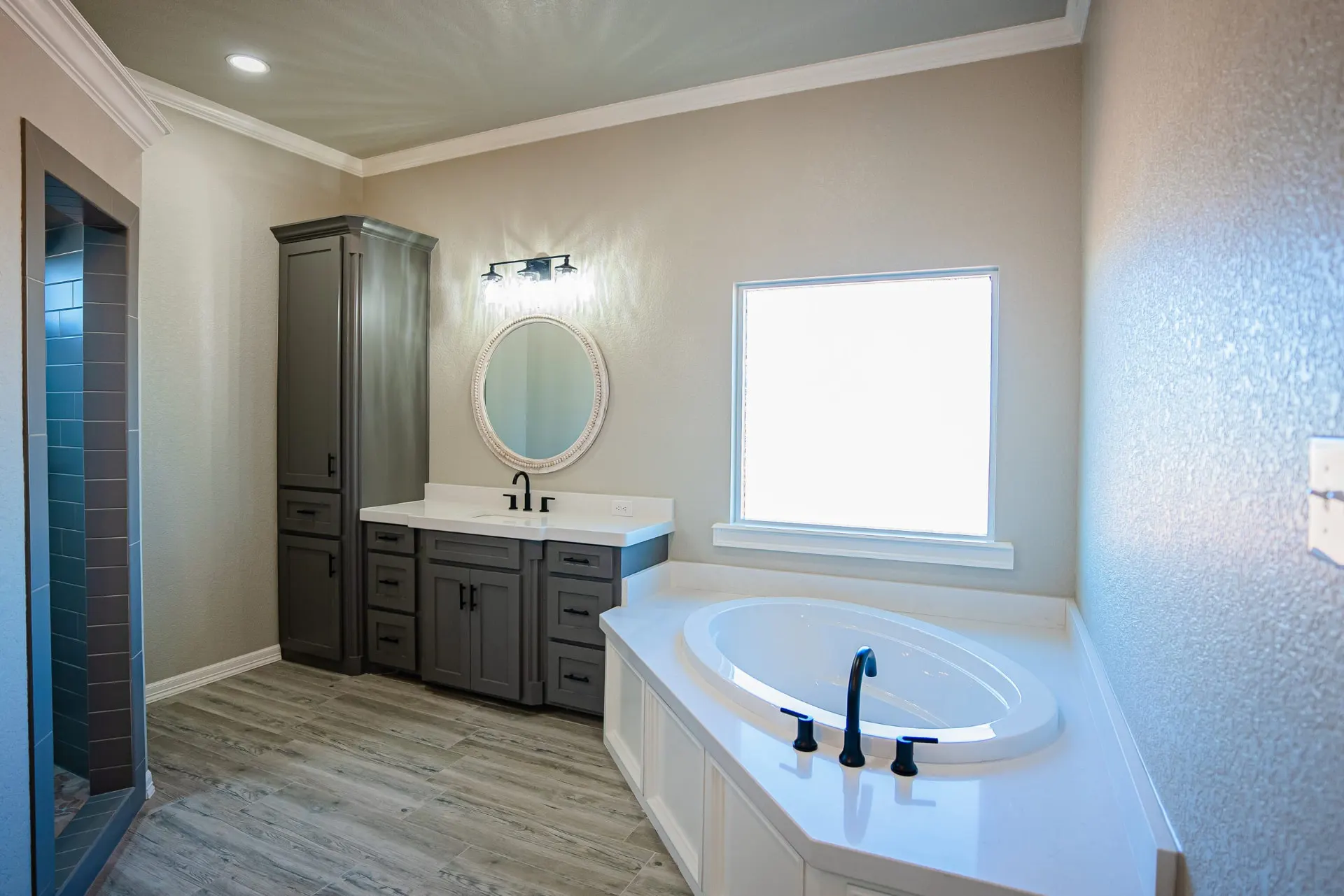 Primary bath at the Eliam St Home with grey cabinetry, a rounded mirror, and a soaking tub under a picture window beside a glass-block shower