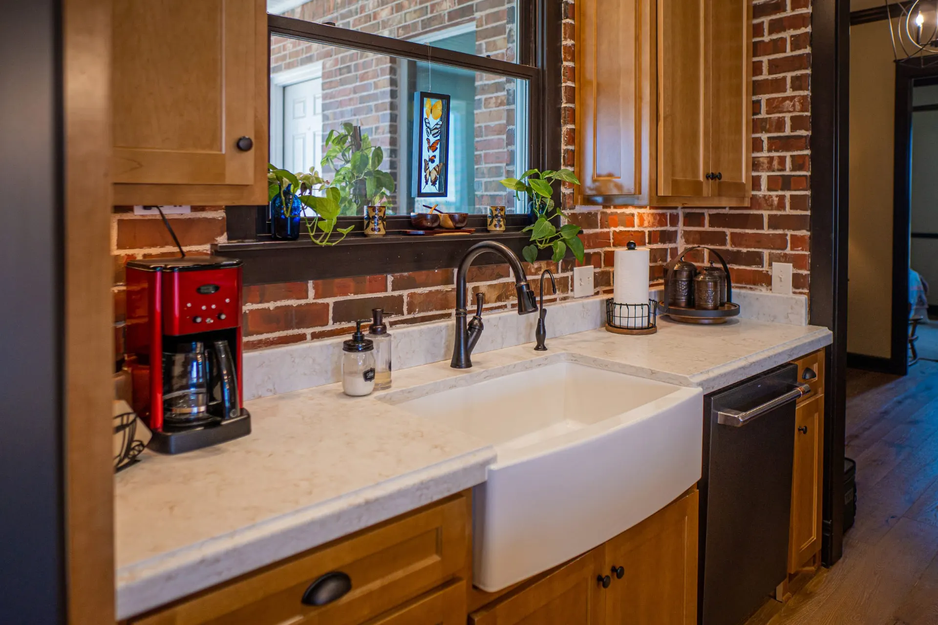Kitchen sink at the Wraparound Porch Home with a white apron-front farmhouse sink and an oil-rubbed bronze gooseneck faucet in natural wood cabinetry, facing a brick-framed interior window