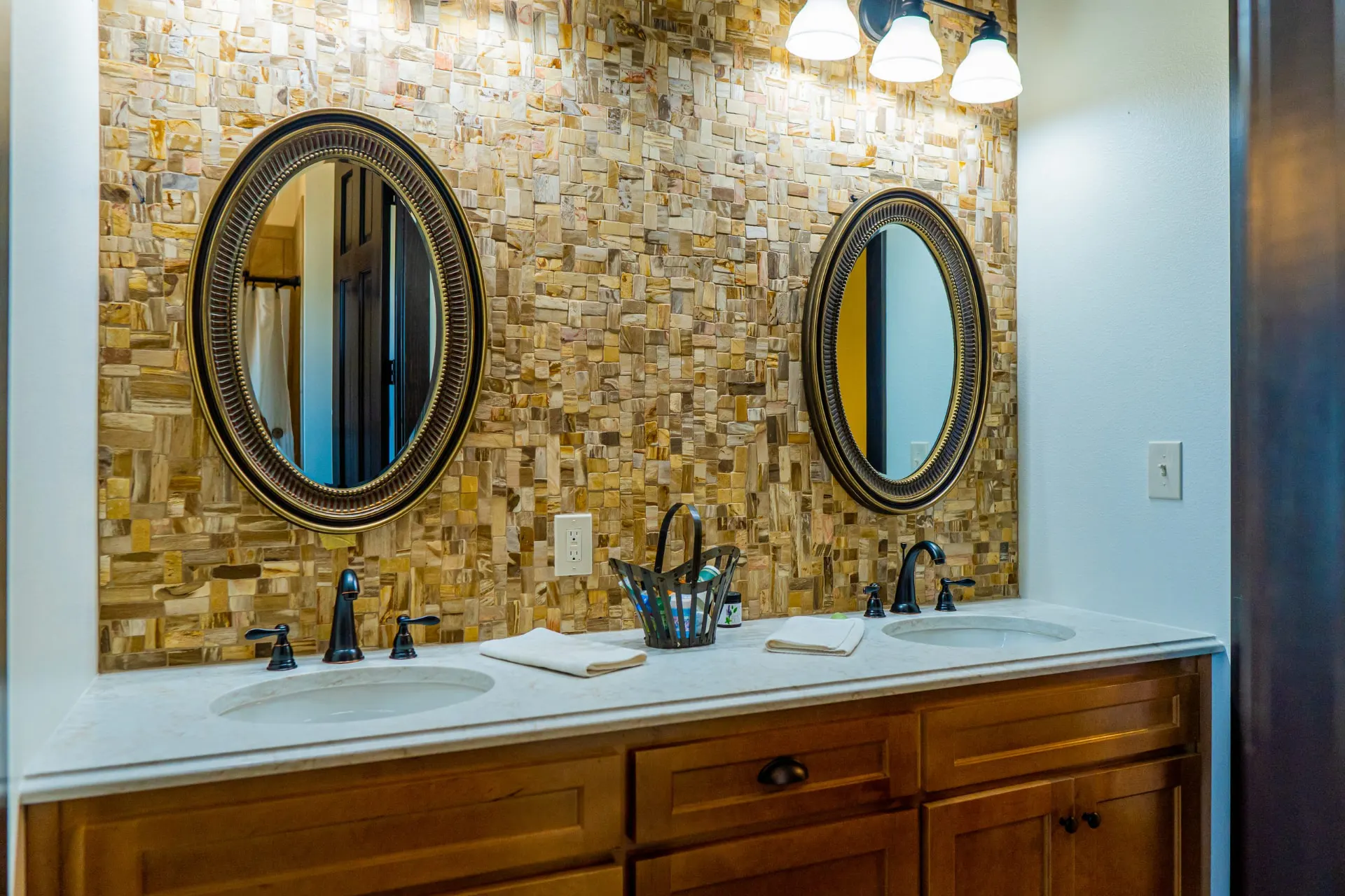 Double vanity in honey-oak cabinetry under twin oval gold-framed mirrors against a mosaic stone tile wall at the Wraparound Porch Home
