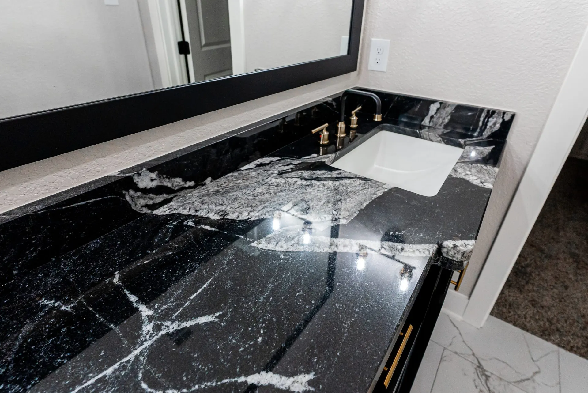 Bathroom vanity at 708 K Ave with a black marble counter and backsplash, white undermount sink, matte black faucet with gold handles and a wide black-framed mirror over marble-look tile