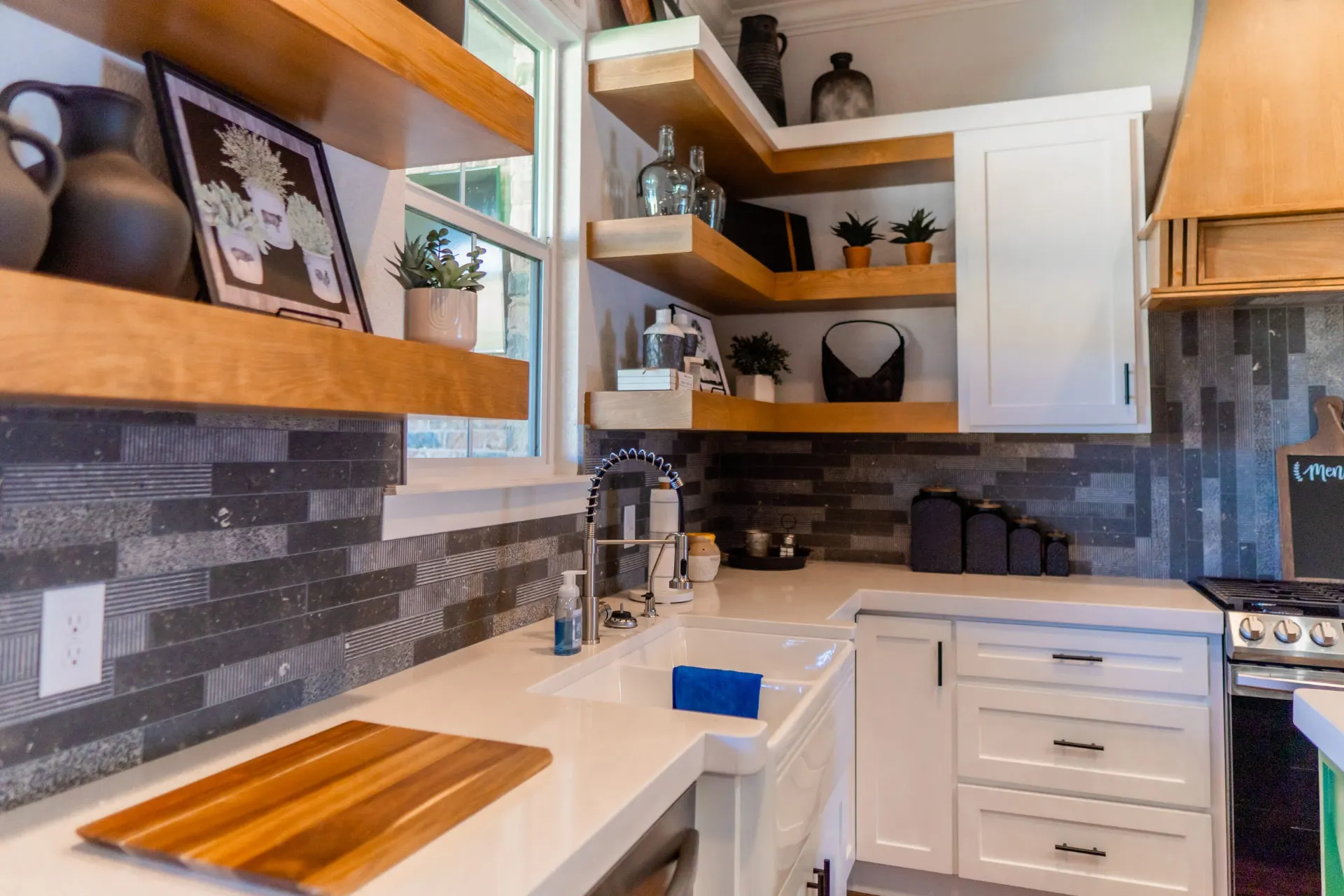 Farmhouse sink detail at the K Ave Farmhouse with a black coil faucet, floating wood shelves, framed art and the dark tiled backsplash