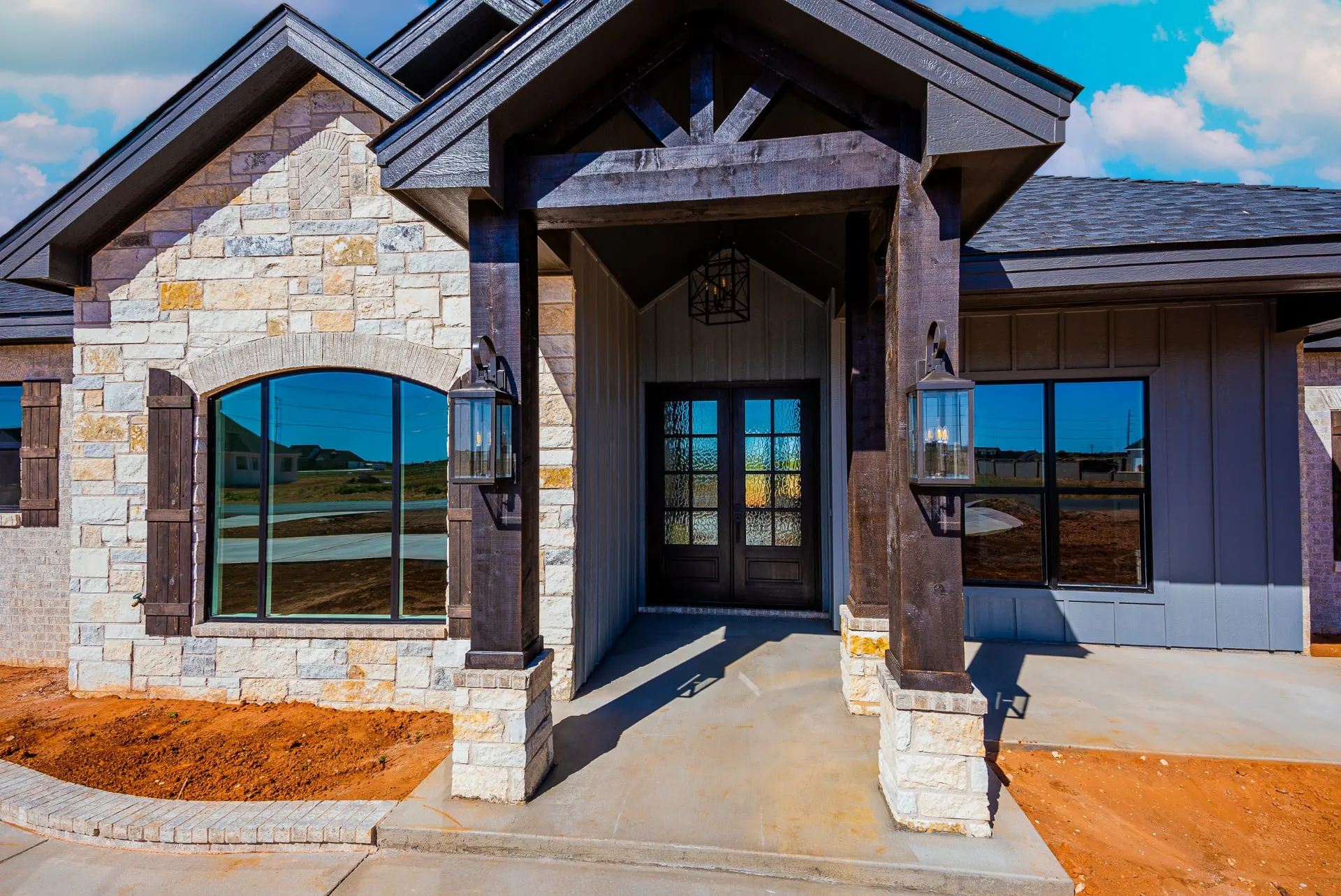 Entry porch of the Eliam St Home under a dark timber gable with lantern sconces on stone bases and dark wood-framed double doors