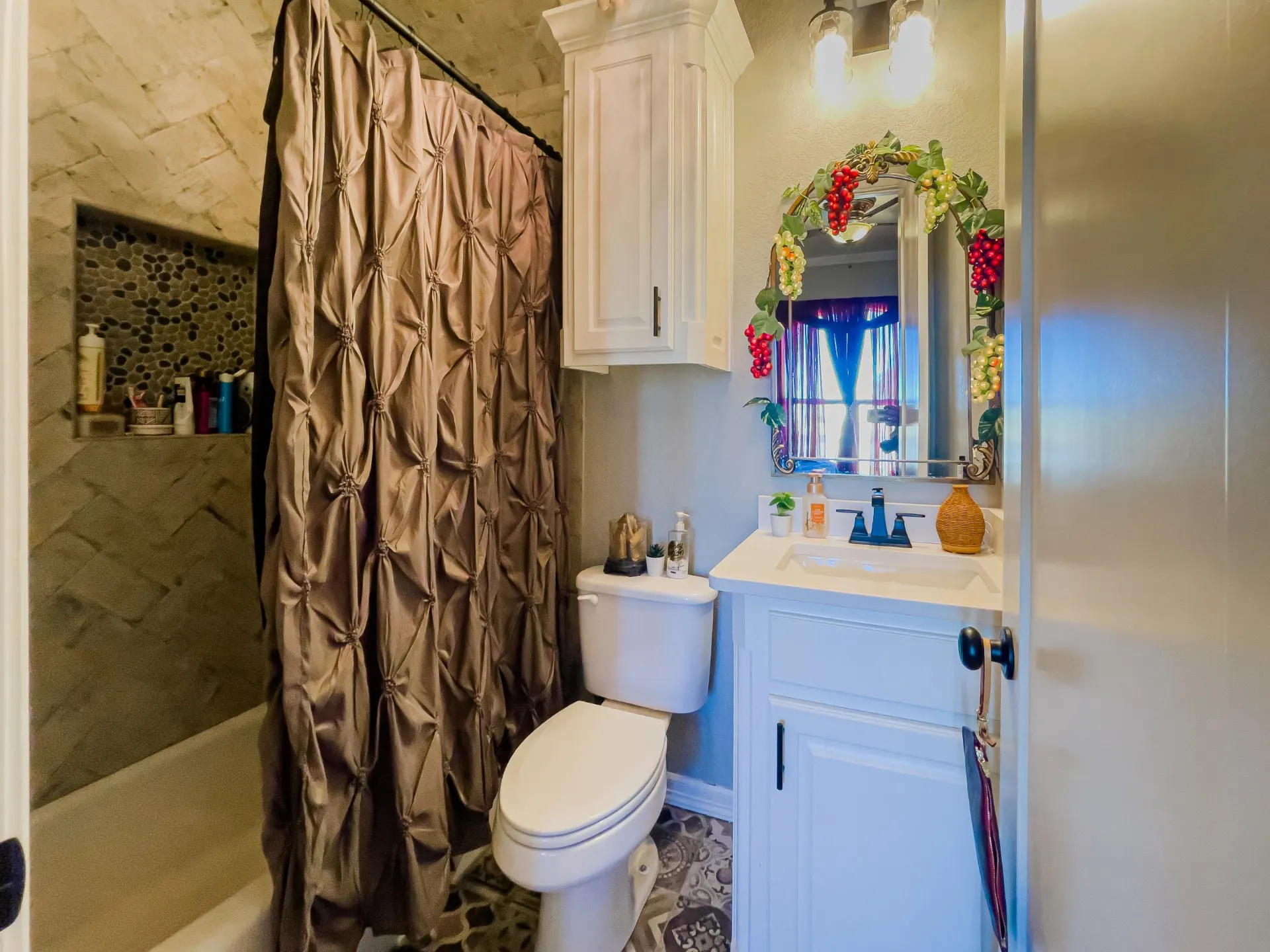 Bath at 8821 Shawn Dr with a white vanity, matte black faucet, an arched mirror framed in grape-vine decor, a taupe shower curtain and patterned floor tile