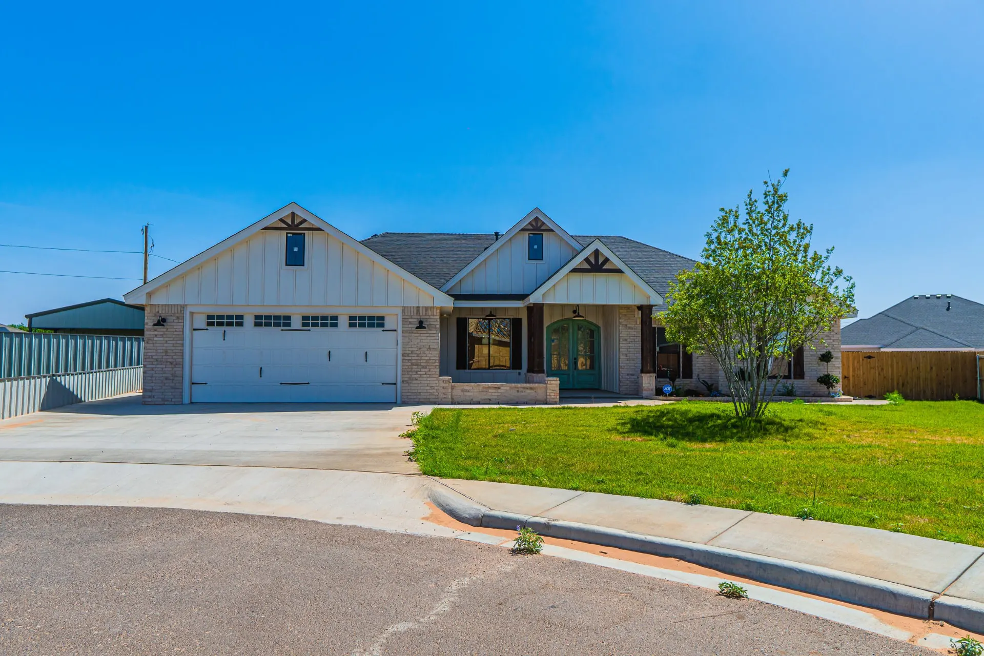 Front exterior of Sarai Home with white board-and-batten gables on light brick, a timber-framed porch with a teal front door, a three-car garage and a young tree in the front lawn
